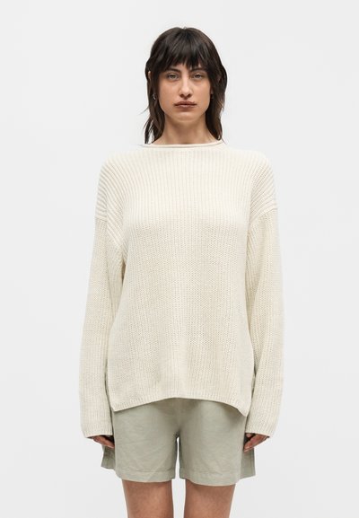 Femme portant un pull crème à maille lâche avec des manches longues et un short beige jusqu'aux genoux, debout devant un fond clair uni.