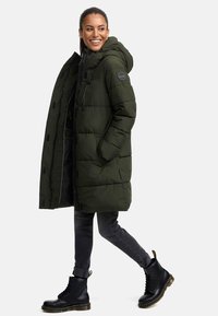 Olive green puffer coat with a hood, featuring zip closure, side pockets, and a quilted design. Worn over dark grey skinny jeans and black boots.