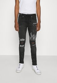 Black denim jeans with distressed detailing, white graphic prints, and a relaxed fit. Features rips at the knees and adjustable waistband.