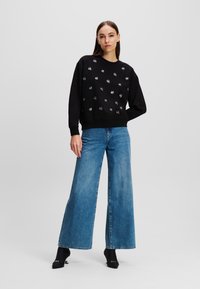 Schwarzer Sweatshirt mit verstreuten weißen und grauen Gesichtsgrafiken, gerippten Bündchen und Saum; kombiniert mit weit geschnittenen blauen Jeans.
