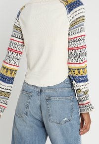 Valkoinen pitkähiha toppi, jossa on kuvioidut neulehihat sinisessä, keltaisessa, punaisessa ja mustassa; yhdistettyinä kulutettuihin vaaleansinisiin farkkuihin.