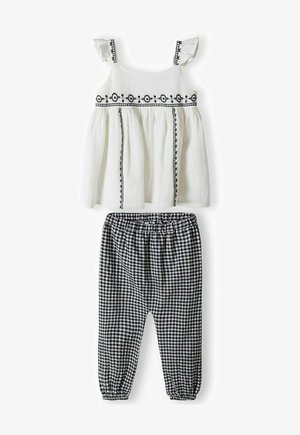 Fehér top fekete hímzett díszítéssel és fodros pántokkal, fekete-fehér kockás nadrággal párosítva, amely elasztikus derékkal rendelkezik.