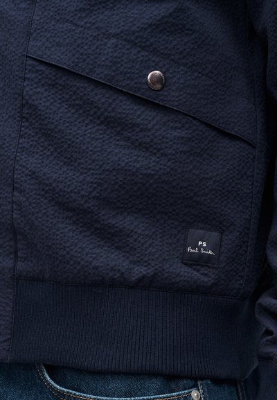 Poche de veste bleu foncé texturée avec un bouton-pression argenté et une petite étiquette noire indiquant "PS Paul Smith" au-dessus de la ceinture côtelée.