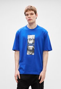 Μπλε βαμβακερό T-shirt με κάθετη γραφική εκτύπω ση μαύρων και λευκών χαρακτήρων και το κείμενο "Jujutsu Kaisen" στο κέντρο. Κοντό μανίκι.