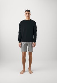 Felpa nera con colletto tondo e maniche lunghe, abbinata a pantaloni corti grigi da ginnastica. Il modello è a piedi nudi su uno sfondo bianco uniforme.
