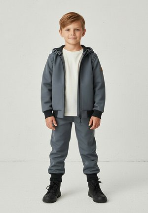 Młody chłopiec stojący, ubrany w szarą bluzę z kapturem, pasujące szare spodnie, białą koszulkę i czarne buty sznurowane na tle gładkiego tła.