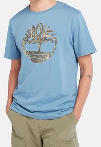 Muž v bledě modrém tričku s logem stromu v maskáčovém vzoru a khaki cargo šortkách, s rukama v kapsách.