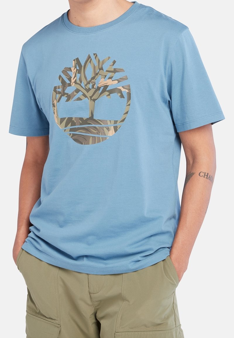 Muž v bledě modrém tričku s logem stromu v maskáčovém vzoru a khaki cargo šortkách, s rukama v kapsách.