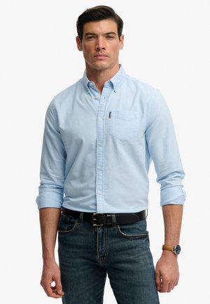 Mężczyzna w jasnoniebieskiej koszuli z długim rękawem podwiniętym do łokcia, ciemnych dżinsach, czarnym pasku i zegarku z brązowym paskiem na lewej ręce.