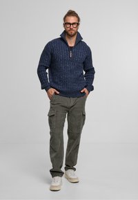 Granatowy sweter z dzianiny z zamkiem przy szyi, wytłaczany wzór, w połączeniu z oliwkowymi spodniami cargo i beżowymi sneakersami wykonanymi z różnych materiałów.