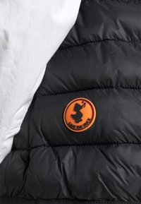 Chaqueta de plumas negra con una sección blanca, un logotipo de goma naranja circular con la silueta de un pato y acolchado tipo colcha para aislamiento.