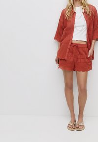 Chemise ample de couleur rouille à tissu texturé, associée à un short assorti avec des bords festonnés et des motifs brodés. Sandales plates.