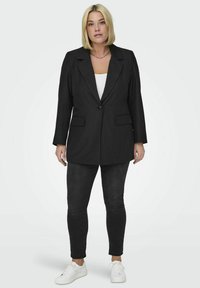 Črn blazer z dolgimi rokavi, prilegajoč kroj, en gumb za zapiranje, dva sprednja žepa, oblečen čez belo majico, v kombinaciji s črnimi kavbojkami in belimi teniskami.