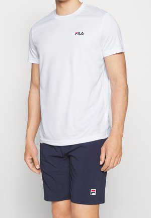 Man wearing white short-sleeve FILA t-shirt and navy FILA shorts, standing with arms relaxed by sides against a plain background.