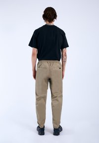 Zwarte T-shirt met korte mouwen, gecombineerd met khaki-elastische broek met twee achterzakken. Model draagt zwarte schoenen, gezien van achteren.