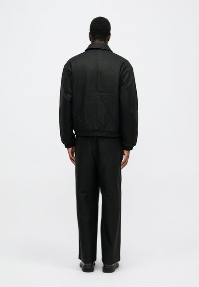 Man staat met zijn rug naar de camera, gekleed in een zwart leren jack, zwarte broek en zwarte schoenen tegen een plain witte achtergrond.