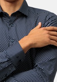 Uomo che indossa una camicia a maniche lunghe blu navy con pois bianchi, con le braccia incrociate e la mano destra appoggiata sul braccio superiore sinistro.