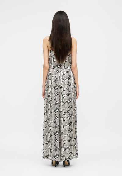 Frau mit langen, dunklen Haaren, die einen ärmellosen, weitbeinigen Jumpsuit mit einem schwarz-weißen Schlangenmuster trägt und mit dem Rücken zur Kamera steht.