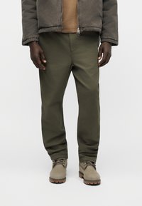 Άνδρας που φοράει χακί παντελόνι, μπεζ σακάκι και μποτάκια με κορδόνια μπέζ, στέκεται μπροστά σε λευκό, απλό φόντο.