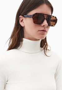 Brązowe okulary przeciwsłoneczne w kształcie tortoise z kanciastym designem i dużymi soczewkami. Rami w subtelny wzór w panterę kontrastują z białym, prążkowanym golfem.