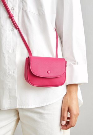 Persona vestida con camisa y pantalones blancos, con un pequeño bolso cruzado de cuero rosa brillante con solapa abotonada.