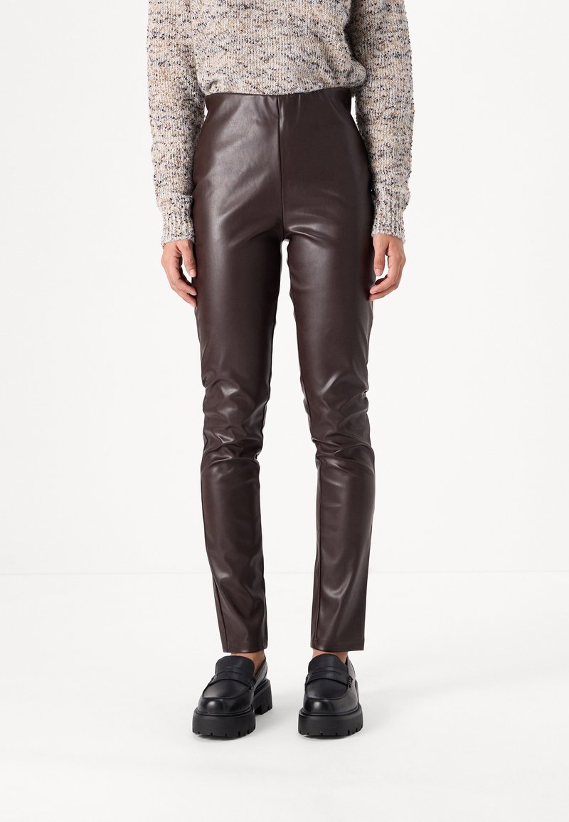 Personne portant un pantalon en cuir marron foncé taille haute, un pull en tricot beige à motifs et des mocassins à plateforme noirs épais, se tenant sur un fond blanc.