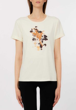 Donna che indossa una t-shirt bianca a maniche corte con un motivo floreale arancione e nero e un design astratto di uccelli sul davanti, abbinata a pantaloni neri.