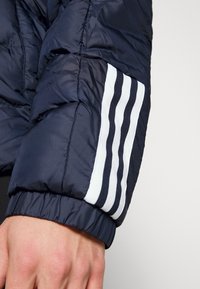 Marineblå puffjakkeærme med tre hvide striber, blank overflade, elastiske manchetter og kviltedetaljer for isolering.