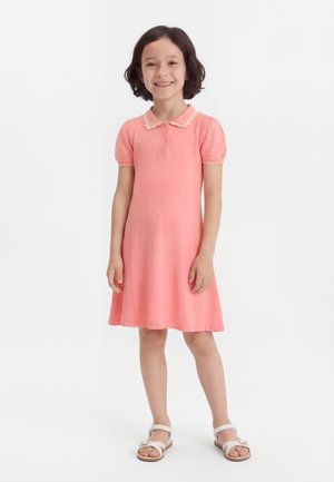 Smilende ung pige med kort mørkt hår iført en koralrosa kortærmet kjole med bølget krave og hvide sandaler på hvid baggrund.