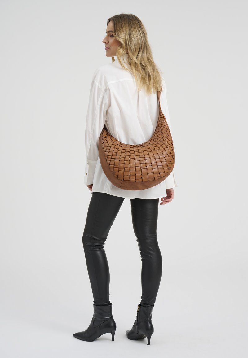 Žena v bílé košili, černých kožených kalhotách a černých botách na podpatku nese velkou hnědou pletenou koženou kabelku přes rameno.