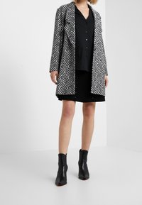 Svart och vit mönstrad lång kappa med geometrisk design, bärs över en svart skjorta med knappar och en svart kjol, i kombination med svarta ankelstövlar.