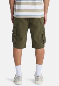 Persoon in olijfgroene cargoshorts, gestreept beige en blauwe shirt, witte enkelkousen en witte sportschoenen, staande met de rug naar voren gericht.