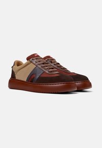 Braune und beige Sneaker aus Wildleder und Canvas, mit runder Zehenpartie, kontrastierenden Texturen und roten Akzenten auf der Zunge und dem Logo.