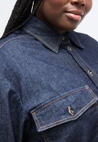 Κοντινό πλάνο ατόμου που φοράει μπλε σκουρόχρωμο τζιν πουκάμισο με κουμπιά, τσέπη στο στήθος και εμφανείς ραφές, αναδεικνύοντας τον γιακά και την υφή του υφάσματος.