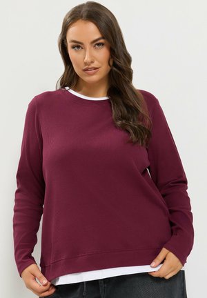 Femme aux longs cheveux bruns portant une chemise à manches longues bordeaux par-dessus un haut blanc, posant devant un fond clair uni.