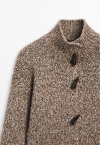 Hnědý pletený svetr s vysokým límcem, který má strukturovaný vzor a tři tmavé knoflíky vpředu. Měkký, silný materiál.