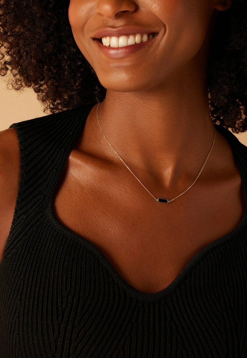 Gouden ketting met een ovale zwarte hanger, gedragen met een zwarte geribbelde tanktop, die de halslijn en de gladde huidtextuur benadrukt.