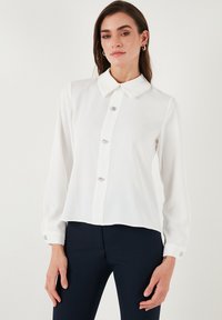 White long-sleeve blouse featuring a classic collar, five metallic buttons, and cuffs with button accents, made from smooth fabric.