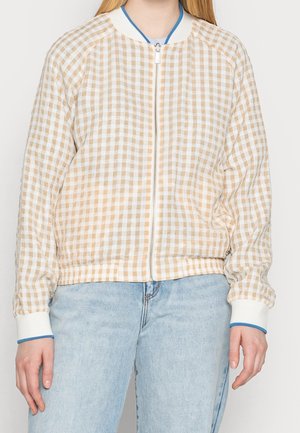 Persoon die een beige-wit geruite jas met ritssluiting draagt, met witte manchetten en lichtblauwe spijkerbroek, staand tegen een egale achtergrond.