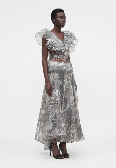 Γυναίκα που στέκεται, φορώντας ένα διαφανές, ασπρόμαυρο floral φόρεμα με μεγάλα βολάν στα μανίκια και μαύρα σανδάλια με ψηλά τακούνια σε λευκό φόντο.