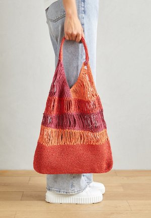 Persoon die een grote, meerkleurige gebreide tas met franjes vasthoudt, draagt een lichte spijkerbroek en witte platform sneakers op een houten vloer.