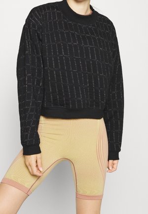 Persoon draagt een zwart sweatshirt met lange mouwen met rasterlijnen en beige geribbelde fietsshorts, staand tegen een effen achtergrond.