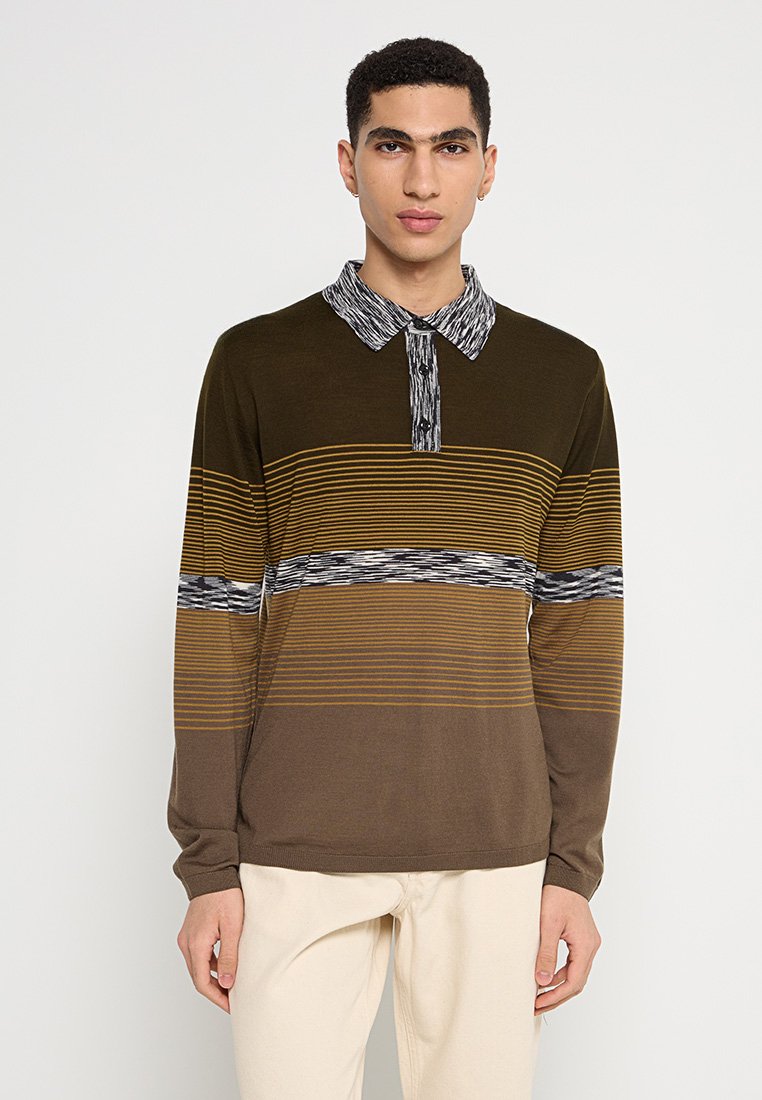 Junger Mann in einem langärmligen, braun gestreiften Poloshirt mit gemustertem Kragen und hellbeigen Hosen, vor einem schlichten Hintergrund stehend.