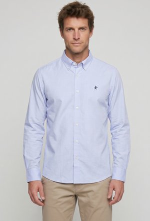 Hombre con camisa azul claro abotonada con un pequeño emblema oscuro en el pecho, combinado con pantalones beige, de pie frente a un fondo liso.