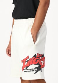 Shorts en coton blanc avec une taille élastique, des poches latérales et un grand motif graphique rouge et noir sur la jambe gauche.