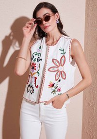Witte mouwloze blouse met kleurrijke bloemenborduursels, ronde halslijn en contrasterende afwerking. Model draagt een zonnebril en witte broek.