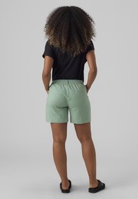 Zielone lniane szorty z elastycznym paskiem i dwoma kieszeniami z tyłu, w zestawie z czarnym topem z krótkim rękawem, widziane od tyłu.