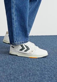 White athletic shoes with gray accents, featuring a textured surface, two dark chevrons, and a contrasting orange detail on the sole.