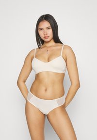 Beige bh og matchende bikini, laget av mykt stoff. Bh-en har tynne stropper og en glatt, sømløs design; bikinitrussen har gjennomsiktige sidelapper.
