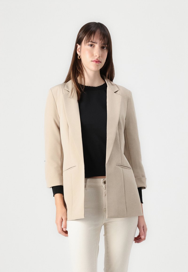 Beige op maat gemaakte blazer met een gelapt kraag, gestructureerde schouders en twee voorzakken, gedragen over een zwart topje. Stevige stofstructuur.